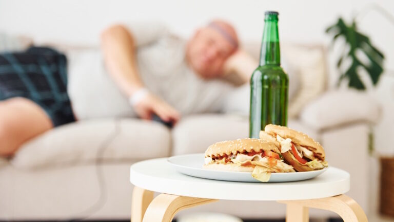 primo piano di un panino con birra, sullo sfondo, fuori fuoco, un uomo sovrappeso sdraiato sul divano