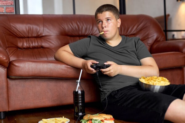 Obesità infantile grave in Italia: oltre 100.000 casi sotto i 17 anni Adolescente sovrappeso, mangia cibo spazzatura mentre gioca ai videogame